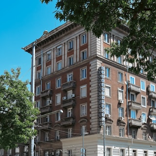 A multi-story brick building with classic architectural details, including ornate balconies and white window frames. The building features a vertical sign reading 'BALKANTOURIST' in blue letters. Surrounding the building, there are leafy green trees, and the sky is clear and blue.