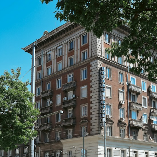 A multi-story brick building with classic architectural details, including ornate balconies and white window frames. The building features a vertical sign reading 'BALKANTOURIST' in blue letters. Surrounding the building, there are leafy green trees, and the sky is clear and blue.