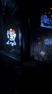 A neon sign that features a stylized face with colorful elements is visible against a dark background. The window showcasing the sign has water droplets on it, suggesting rain or condensation. In the distance, blurry images of a television screen or brightly lit area can be seen through the glass, alongside some twinkling lights.