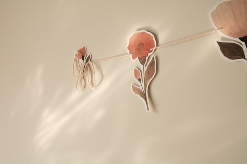 A delicate garland of paper flowers is hung on a neutral-colored wall, casting soft shadows. The flowers are in muted tones of pink and brown with simple leafy stems.