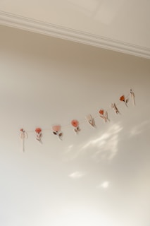 A series of delicate, pastel-colored floral cutouts hang from a string against a soft cream-colored wall. The arrangement consists of simple flower shapes in muted pinks and oranges, casting subtle shadows that create a gentle, artistic display.