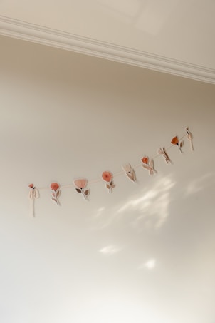 A series of delicate, pastel-colored floral cutouts hang from a string against a soft cream-colored wall. The arrangement consists of simple flower shapes in muted pinks and oranges, casting subtle shadows that create a gentle, artistic display.