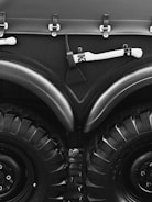 A close-up view of a large vehicle's wheels and bodywork, featuring thick black tires with deep treads. Above the wheels, there is a matte black surface with metal fasteners and a white-handled tool, possibly an axe, mounted horizontally.