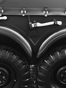 A close-up view of a large vehicle's wheels and bodywork, featuring thick black tires with deep treads. Above the wheels, there is a matte black surface with metal fasteners and a white-handled tool, possibly an axe, mounted horizontally.