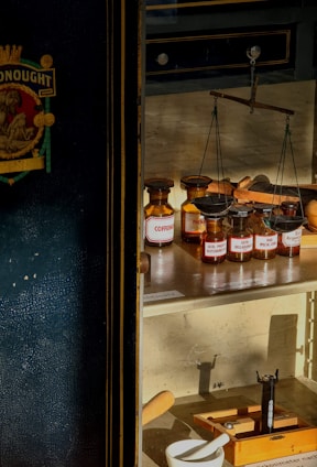 A vintage display features a collection of apothecary jars with labeled contents such as 'Coffein,' 'Acid. Phenylbarbital,' and 'Extr. Belladonna.' A set of balanced scales is positioned among the jars, indicating a historical or scientific context. The scene is warmly lit by sunlight, casting distinct shadows within the display case. The surrounding area suggests a museum or retro pharmacy setting with elements like a mortar and pestle and wooden equipment.