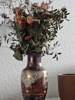 A decorative vase with intricate patterns and designs, holding a bouquet of dried flowers. The vase features ornate gold detailing and a painted portrait. Beside the vase is a simple brown cup. The background is a textured white wall.