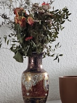 A decorative vase with intricate patterns and designs, holding a bouquet of dried flowers. The vase features ornate gold detailing and a painted portrait. Beside the vase is a simple brown cup. The background is a textured white wall.
