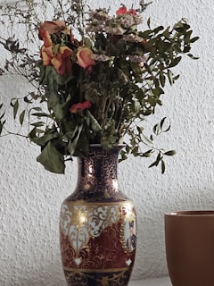 A decorative vase with intricate patterns and designs, holding a bouquet of dried flowers. The vase features ornate gold detailing and a painted portrait. Beside the vase is a simple brown cup. The background is a textured white wall.