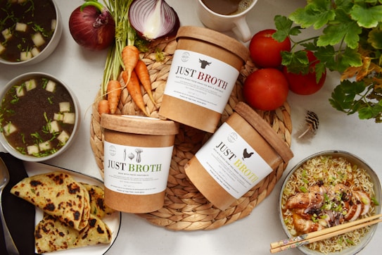 A variety of ingredients and dishes are artfully arranged on a light-colored surface. In the center, there are three containers labeled 'Just Broth' placed on a woven mat, surrounded by fresh vegetables like carrots, onions, and tomatoes. To the left, two bowls of soup with green garnishes and diced white pieces are placed. Additionally, there is a plate of sliced flatbreads and on the right, a bowl of seasoned noodles topped with roasted chicken and garnished with green onions. A cup of coffee is partially visible at the top.