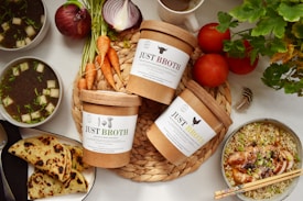 A variety of ingredients and dishes are artfully arranged on a light-colored surface. In the center, there are three containers labeled 'Just Broth' placed on a woven mat, surrounded by fresh vegetables like carrots, onions, and tomatoes. To the left, two bowls of soup with green garnishes and diced white pieces are placed. Additionally, there is a plate of sliced flatbreads and on the right, a bowl of seasoned noodles topped with roasted chicken and garnished with green onions. A cup of coffee is partially visible at the top.