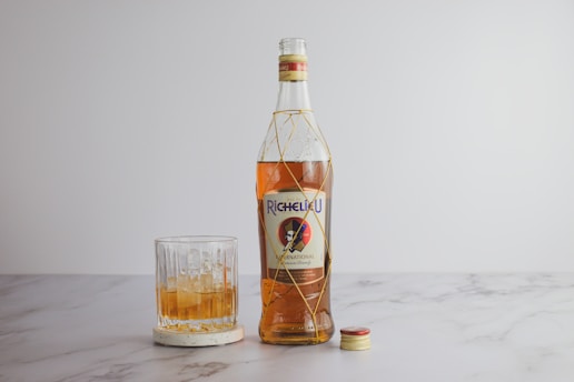 A bottle of Richelieu International brandy stands next to a glass filled with a similar amber liquid and ice, placed on a marble surface. The golden bottle cap is off and positioned on the right side of the bottle.
