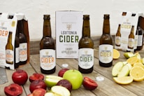 Several bottles of Loxtonia Cider are arranged on a rustic wooden table, accompanied by sliced red and green apples, oranges, and nectarines. The cider bottles have labels indicating different varieties, including crispy apple and sparkling apple. Packaging boxes with the Loxtonia brand are also present in the background.