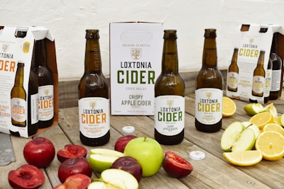 Several bottles of Loxtonia Cider are arranged on a rustic wooden table, accompanied by sliced red and green apples, oranges, and nectarines. The cider bottles have labels indicating different varieties, including crispy apple and sparkling apple. Packaging boxes with the Loxtonia brand are also present in the background.