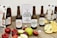 Several bottles of Loxtonia Cider are arranged on a rustic wooden table, accompanied by sliced red and green apples, oranges, and nectarines. The cider bottles have labels indicating different varieties, including crispy apple and sparkling apple. Packaging boxes with the Loxtonia brand are also present in the background.