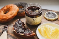 A wooden board holds a jar of blackcurrant and orange jam with cinnamon flavor. Beside the jar, there is a sliced piece of bread, spread with the same dark, glossy jam. Two bagels are placed nearby, one plain and the other coated with poppy seeds. A small plate with neatly arranged pats of butter and a butter knife with some jam on it are also visible.
