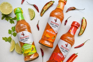 Three bottles of Nando's Peri-Peri sauce are arranged on a light surface, surrounded by sliced limes, red chili peppers, and green herbs. The bottles are labeled as Lemon & Herb Extra Mild, Medium, and Hot, each with distinctive colored labels reflecting their flavor intensity.