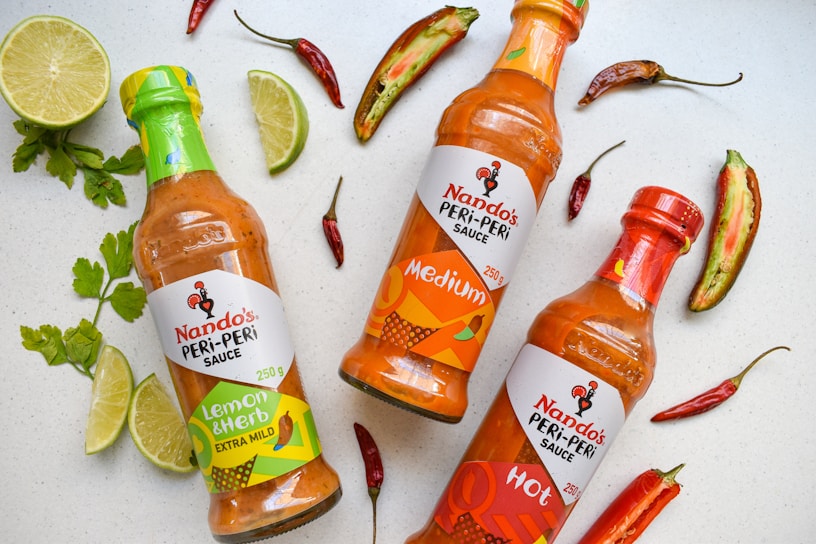 Three bottles of Nando's Peri-Peri sauce are arranged on a light surface, surrounded by sliced limes, red chili peppers, and green herbs. The bottles are labeled as Lemon & Herb Extra Mild, Medium, and Hot, each with distinctive colored labels reflecting their flavor intensity.