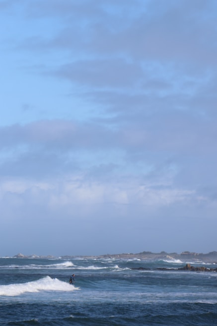 A person is surfing on small waves in an expansive ocean setting with a distant shoreline. The sky is partly cloudy, and the sea is a deep blue color, creating a contrast with the white surf. The scene conveys a sense of adventure and tranquility.