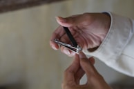 A person is using a metal nail clipper to trim their fingernails. The hand holding the clipper wears a light-colored sleeve, while the other hand is bare, with fingers positioned to clip the nail.