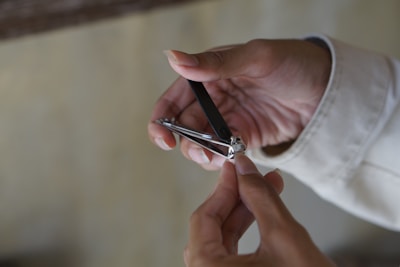 A person is using a metal nail clipper to trim their fingernails. The hand holding the clipper wears a light-colored sleeve, while the other hand is bare, with fingers positioned to clip the nail.