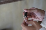 A person's hands are engaged in cutting fingernails using a metallic nail clipper. The fingers are delicately positioned, and the lighting is soft, highlighting the skin tones and the metallic sheen of the clipper.