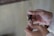 A person's hands are engaged in cutting fingernails using a metallic nail clipper. The fingers are delicately positioned, and the lighting is soft, highlighting the skin tones and the metallic sheen of the clipper.