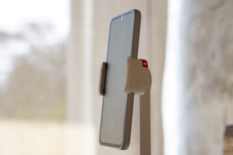 A mobile phone mounted on a minimalist stand with a neutral-colored background. The phone is held securely with a clamp that has a visible red accent. The background is softly blurred, creating a sense of depth.