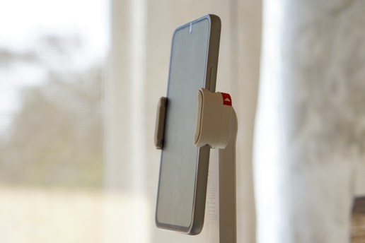 A mobile phone mounted on a minimalist stand with a neutral-colored background. The phone is held securely with a clamp that has a visible red accent. The background is softly blurred, creating a sense of depth.