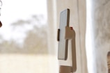 A smartphone mounted on a robotic arm-like holder, positioned vertically. In the background, there are sheer curtains allowing diffused natural light into the room. Part of a person's face is visible on the left edge of the image, suggesting interaction with the phone.