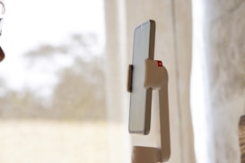 A smartphone mounted on a robotic arm-like holder, positioned vertically. In the background, there are sheer curtains allowing diffused natural light into the room. Part of a person's face is visible on the left edge of the image, suggesting interaction with the phone.
