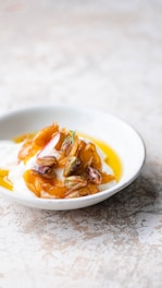 A bowl filled with a creamy white substance, likely yogurt, topped with golden caramelized pieces, pistachios, and a hint of herb. The dish is presented on a light-colored, textured surface.