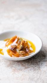 A bowl filled with a creamy white substance, likely yogurt, topped with golden caramelized pieces, pistachios, and a hint of herb. The dish is presented on a light-colored, textured surface.