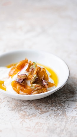 A bowl filled with a creamy white substance, likely yogurt, topped with golden caramelized pieces, pistachios, and a hint of herb. The dish is presented on a light-colored, textured surface.
