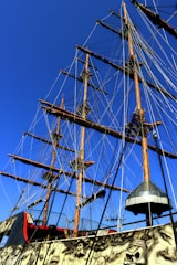 Tall ship masts with numerous ropes and rigging against a clear blue sky. The ship has a decorative exterior with a pirate theme, featuring intricate details like skulls.