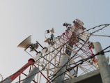 A tall metal communication tower extends upward against the backdrop of a clear sky. The structure is equipped with several dish antennas and various electronic components, featuring a lattice framework painted in red and white for high visibility. Barbed wires are present in the foreground, suggesting a secured area.