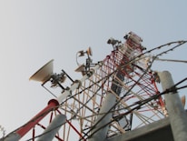 A tall metal communication tower extends upward against the backdrop of a clear sky. The structure is equipped with several dish antennas and various electronic components, featuring a lattice framework painted in red and white for high visibility. Barbed wires are present in the foreground, suggesting a secured area.