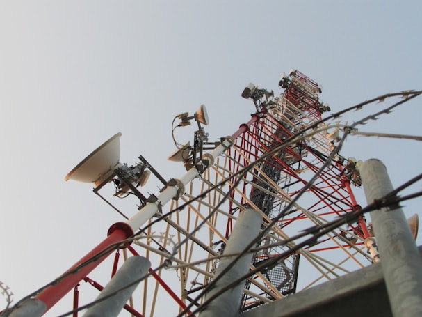 A tall metal communication tower extends upward against the backdrop of a clear sky. The structure is equipped with several dish antennas and various electronic components, featuring a lattice framework painted in red and white for high visibility. Barbed wires are present in the foreground, suggesting a secured area.