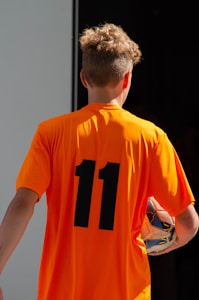 A person is wearing an orange sports jersey with the number 11 on the back, holding a soccer ball under their arm. The individual has short, curly hair and appears to be in a sports facility or outdoor setting.