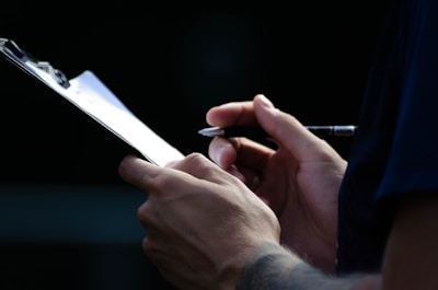 A close-up of a person holding a pen and a clipboard, seemingly taking notes or writing something. The setting appears to be dimly lit with focus on the hands and the writing action.