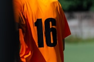 A vibrant orange football jersey displayed on a hanger with a blurred stadium backdrop.