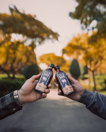 Two hands holding bottles of cold brew coffee together against a background of vibrant autumn foliage. The bottles are dark with eye-catching labels, and the scene conveys a sense of camaraderie and relaxation in a natural setting.