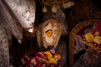 The image features a traditional handmade mask with a painted face, surrounded by ornate, decorative items. The mask has distinct features with bright colors and intricate details, set against a backdrop of vibrant textiles and embellishments such as tassels and embroidery.
