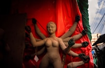 A clay sculpture of a goddess with multiple arms is being crafted, set against a bright red backdrop. The sculpture is partially completed, with visible cracks and raw material edges. The scene is illuminated with natural light, casting shadows that add depth to the figure. Surrounding elements include cloth and wires, indicating an ongoing work in progress.