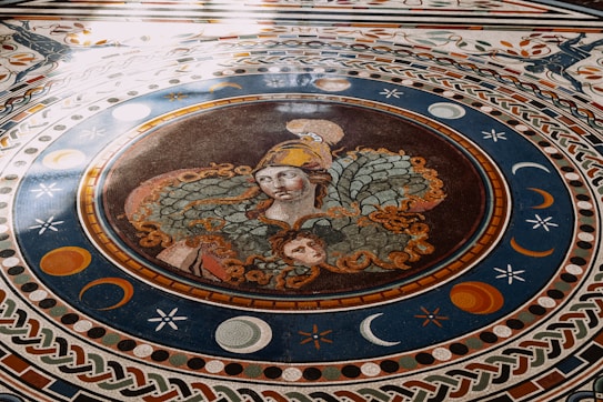 A detailed mosaic on a floor depicting a mythological scene with a central figure wearing a helmet adorned with intricate designs surrounded by celestial motifs like moons and stars. The mosaic features vibrant colors and elaborate patterns combining circular and geometric borders.