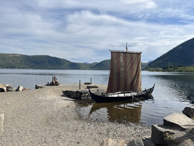 A Viking-style longboat with a striped sail is moored by a rocky shore, with a person sitting on the rocks near the water. The background features a serene lake surrounded by rolling green hills and a cloudy sky.