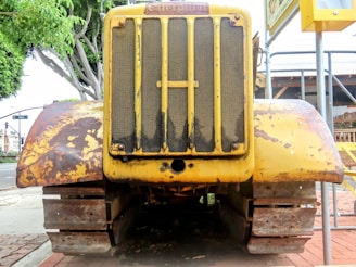 A vintage Caterpillar bulldozer with a weathered yellow finish is positioned in a stationary stance. The machinery features a prominent front grille with rust spots and wears heavy-duty tracks. Surrounding the scene are elements like trees, sidewalks, and signs, hinting at an urban setting.