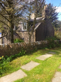 A quaint stone cottage partially obscured by trees, with a sloped roof and several chimney stacks. A stone boundary wall runs along a neatly maintained garden with stepping stones on a grassy path. Lush greenery and tall trees frame the scene under a clear blue sky.