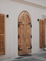 A detailed wooden double door with intricate paneling set in an arched frame. The door is flanked by two wooden window shutters mounted on the exterior wall. The surrounding area has a smooth light-colored wall with two small black wall-mounted lights. The ground is partially visible with a small door mat placed in front of the door.