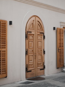 A detailed wooden double door with intricate paneling set in an arched frame. The door is flanked by two wooden window shutters mounted on the exterior wall. The surrounding area has a smooth light-colored wall with two small black wall-mounted lights. The ground is partially visible with a small door mat placed in front of the door.