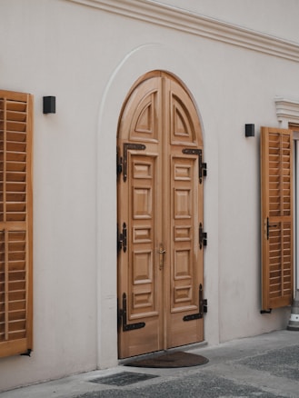 A detailed wooden double door with intricate paneling set in an arched frame. The door is flanked by two wooden window shutters mounted on the exterior wall. The surrounding area has a smooth light-colored wall with two small black wall-mounted lights. The ground is partially visible with a small door mat placed in front of the door.